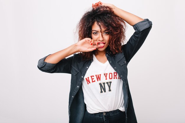 Close-up portrait of sensual mulatto woman in denim jacket isolated. Indoor photo of curly brunette girl playfully posing and ites finger.