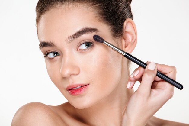 Close up portrait of sensual woman with clean shining skin applying eye shadow using brush