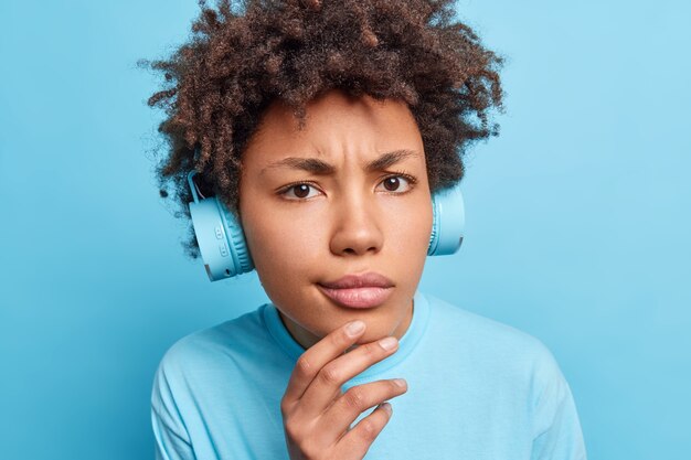 Close up portrait of serious Afro American girl keeps hand on chin looks displeased  dressed casually listens music via headphones or learns new foreign words isolated over blue wall