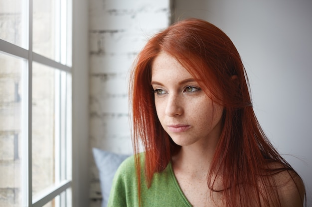 Close up portrait of serious beautiful young european female with freckles and ginger loose hair sitting on windowsill