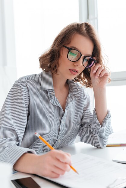 Close-up portrait of serious brunette woman touching her glasses while working  with papers at home