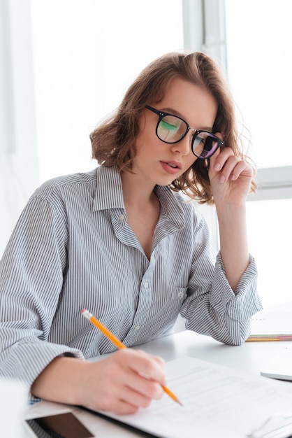 Close-up portrait of serious brunette woman touching her glasses while working  with papers at home