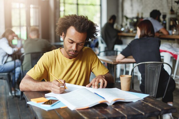 Close-up portrait of serious dark-skinned male student wearing yellow T-shirt sitting at cafe during break drinking coffee and preparing for lessons writing in copybook from book with pencil
