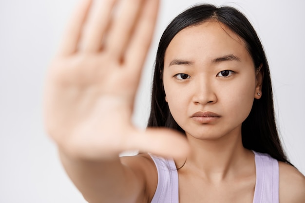 Close up portrait of serious korean woman stretch out hand, showing stop sign with confident face expression, rejecting something, saying no, standing on white