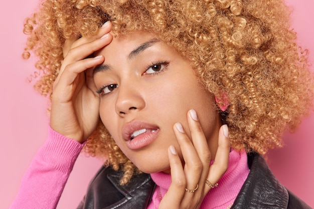 Close up portrait of serious lovely young woman with curly hair natural beauty touches face gently looks directly at camera has healthy skin well cared complexion isolated over pink background.