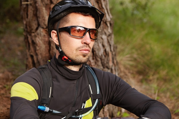 Free photo close up portrait of serious and thoughtful rider with stubble wearing cycling clothing, protective helmet and glasses sitting outdoors at tree and looking ahead of him, thinking about his life