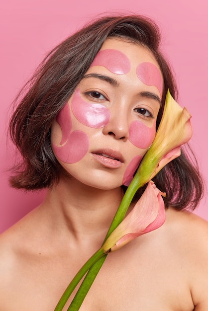 Close up portrait of serious young Asian woman with short dark hair applies hydrogel patches on face to refresh skin holds flower stands naked against pink wall undergoes beauty procedures.