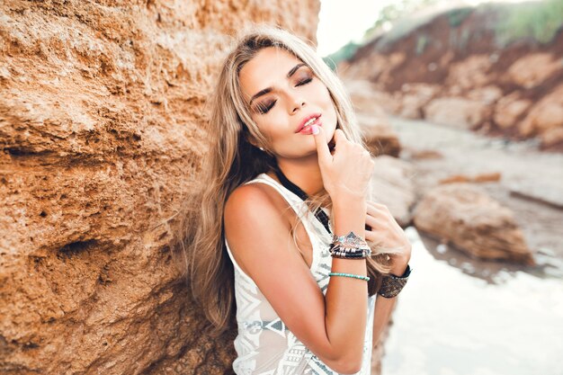 Close-up portrait of sexy blonde girl with long hair posing to the camera on stone background. She keeps finger on lips and eyes closed.