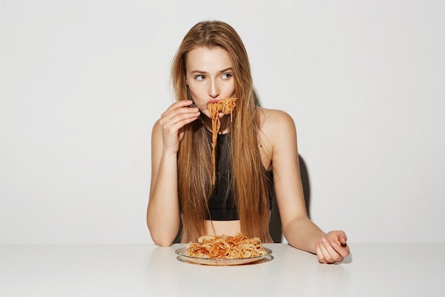 Крупным планом портрет сексуальная блондинка с длинными волосами, сидя за столом, есть спагетти, глядя в сторону с расслабленным и кокетливым выражением.