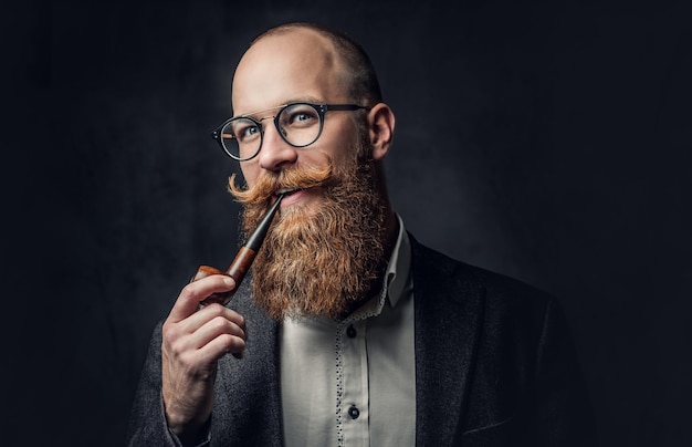 Close up portrait of shaved head aristocratic male in eyeglasses smoking pipe over grey background.