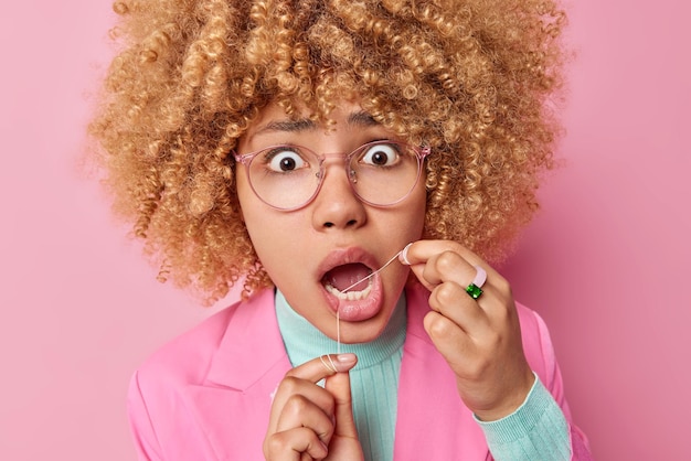 Foto gratuita ritratto ravvicinato di una giovane donna dai capelli ricci scioccata che usa il filo interdentale i suoi denti fissa gli occhi infastiditi ha imbarazzato l'espressione facciale indossa abiti formali pone interni cura dei denti e concetto di igiene orale