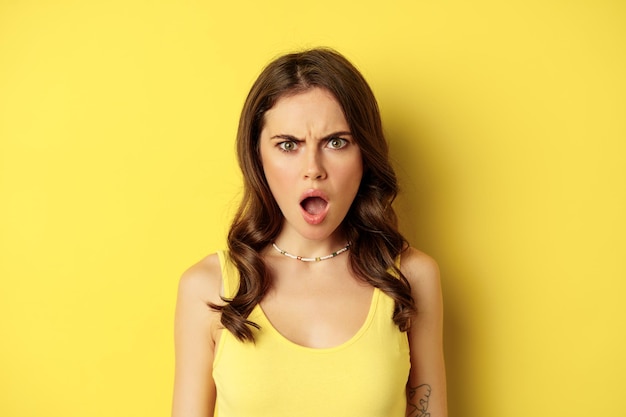 Close up portrait of shocked young woman drop jaw, gasping and looking frustrated, standing against yellow background