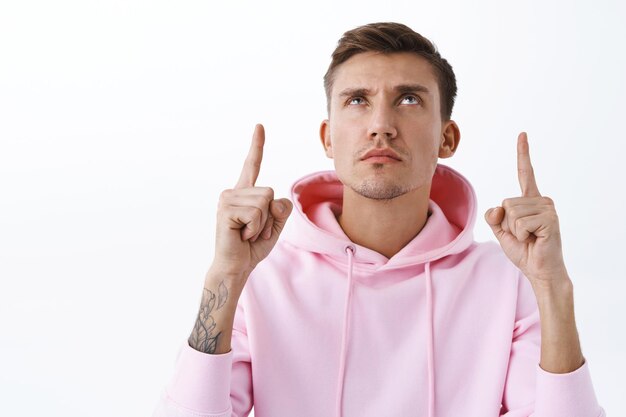 Close-up portrait of skeptical, serious-looking blond man in pink hoodie, frowning, pointing fingers and looking up with suspicious look, express disbelief, standing white wall