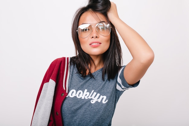 Close-up portrait of smart brunette lady wears glasses posing with hand up. Attractive european girl in trendy t-shirt playing with shiny hair isolated.