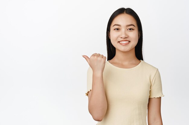 Close up portrait of smiling asian woman pointing left looking happy demonstrating sale info promo text on copy space white background