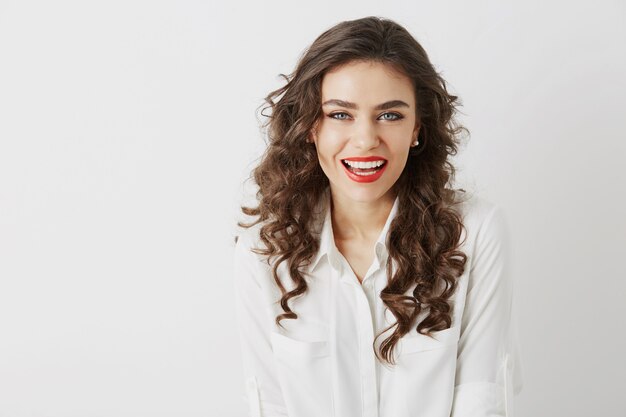 Close-up portrait of smiling attractive woman with white teeth, long curly hair, red lipstick make-up  isolated wearing white blouse