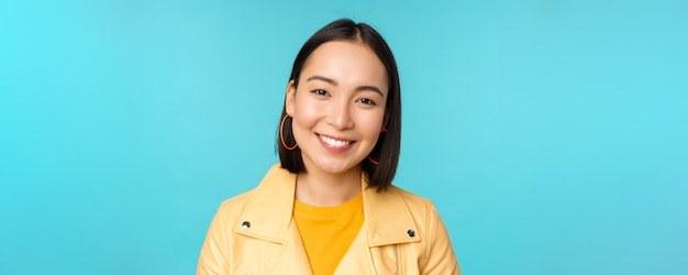 Close up portrait of smiling beautiful asian woman with white teeth looking happy at camera posing in yellow jacket over blue studio background