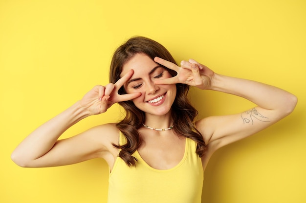 Close up portrait of smiling beautiful girl, showing peace v-sign and looking happy, posing for summer photo, standing against yellow background.