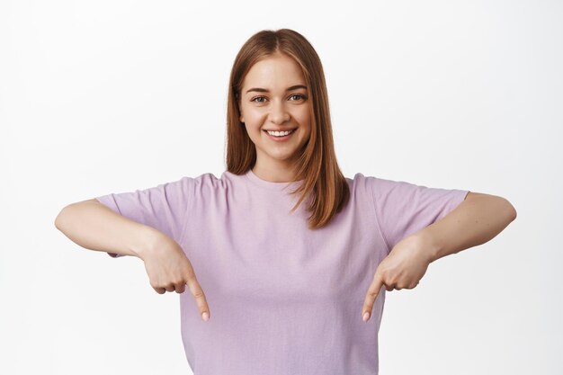 Close up portrait of smiling blond girl pointing fingers down, looking happy and confident, showing advertisement below, store discounts banner, white background. Copy space