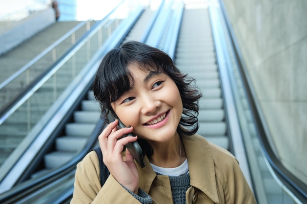 웃는 갈색 머리 여자 한국 여자의 초상화를 닫습니다 에스컬레이터 내려 전화 통화 회담 t