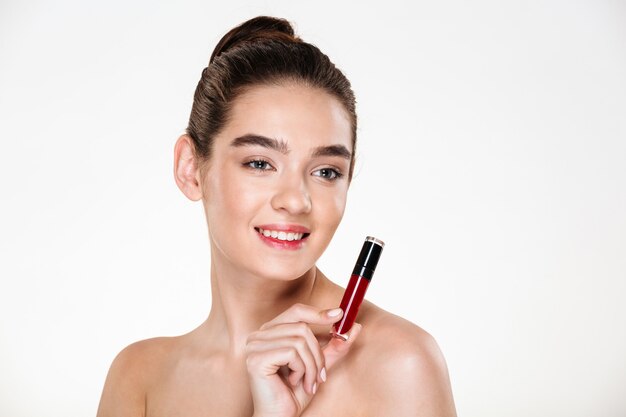 Close up portrait of smiling brunette woman wearing makeup holding lip gloss at her face and looking aside