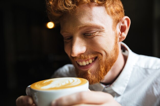 Close-up portrait of smiling curly redhead bearded man tasting coffee in cup