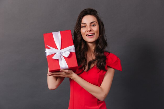 Close-up portrait of smiling dark-haired woman holding decorated box with gift. Lady in red is smiling sincerely on black background.