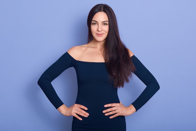 Free photo close up portrait of smiling elegance woman wearing stylish dress standing with hands on hip and looking smiling at camera, winsome girl posing with bare shoulders isoalted on blue.