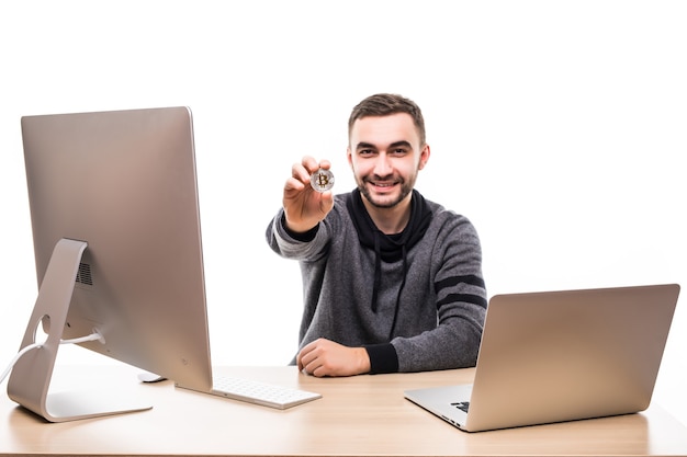 Close up portrait of a smiling entrepreneur holding bitcoin while sitting at desk with laptop and computer isolated over white