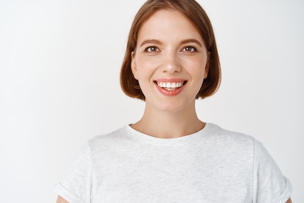 Free photo close-up portrait of smiling girl with short hair and t-shirt, looking cheerful . woman with hopeful eyes standing against white wall