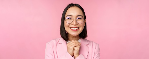 Close up portrait of smiling happy businesswoman in glasses clench hands together thankful excited of smth begging or say please standing over pink background