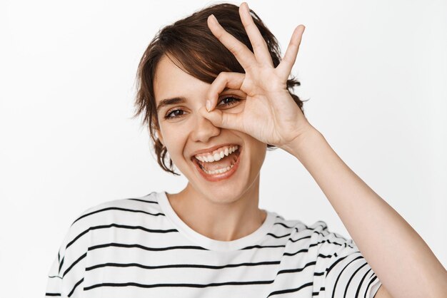 Close up portrait of smiling happy woman showing ok okay sign and looking satisfied recommending something good praising product standing over white background