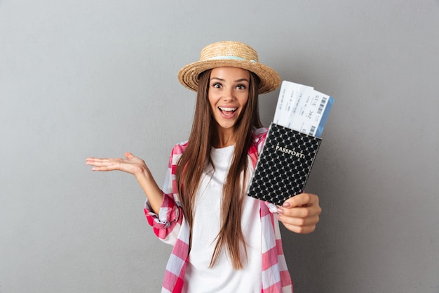 Close up portrait of a smiling happy woman traveller in straw hat showing passport with plane tickets