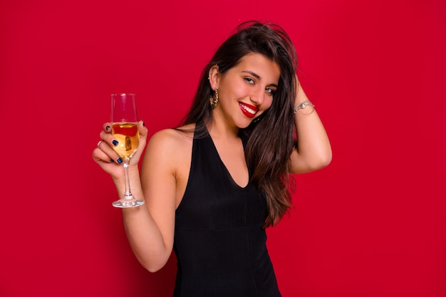 Free photo close-up portrait of smiling happy woman with long dark hair posing with a glass of champagne over isolated red background