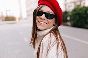 Close up portrait of smiling lovely girl with long light brown hair wearing sunglasses and red beret is looking at camera and laughing