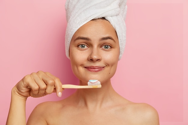 Close up portrait of smiling optimistic woman with fresh skin, holds toothbrush with toothpaste, wears white towel on head, looks directly, has oral hygiene procedure