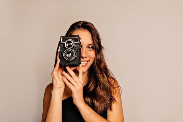 Close up portrait of smiling pretty woman with long curly hair and bright make up posing with retro camera on beige