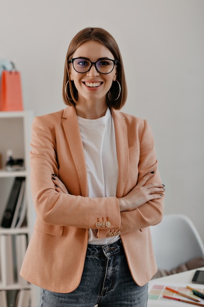 Foto gratuita ritratto del primo piano della donna sorridente di affari dai capelli corti in maglietta bianca in posa con le braccia incrociate in ufficio bianco.