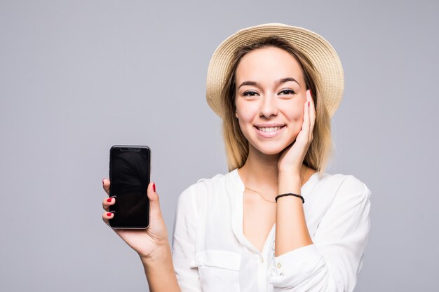 Close up portrait of a smiling woman showing blank screen mobile phone while standing isolated over gray wall