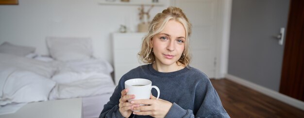 Close up portrait of smiling young blond woman sits with cup of tea in bedroom rests at home enjoys