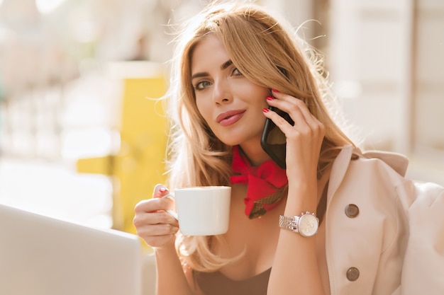 Close-up portrait of spectacular blonde woman looking to camera with interest while talking on phone