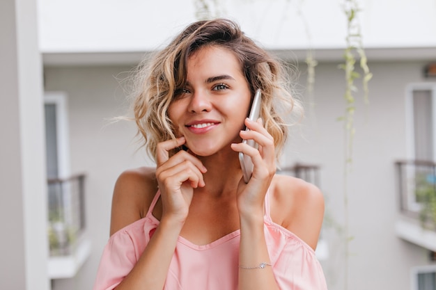 Close-up portrait of spectacular young woman in pink blouse touching her face during phone conversation. Cute blonde girl calling friend from hotel balcony.