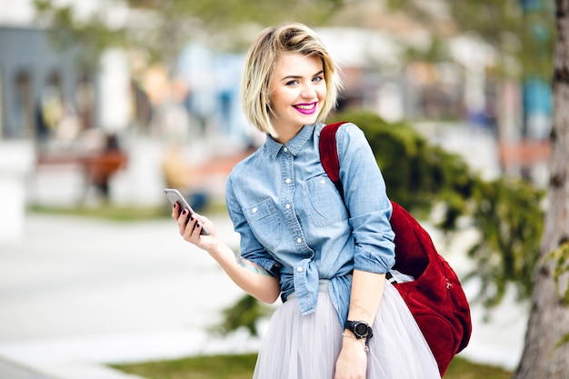 Un ritratto del primo piano di una ragazza sorridente in piedi con i capelli biondi corti e le labbra rosa brillante che hoding uno smartphone