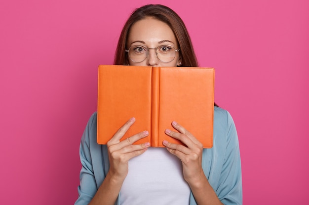 Free photo close up portrait of student girl hides behind book, studio photo over pink