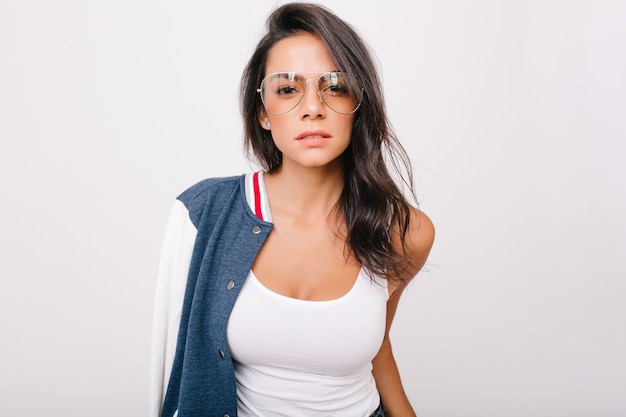 Close-up portrait of stunning dark-haired girl in white tank-top looking with interest. Indoor photo of adorable european lady in stylish glasses chilling