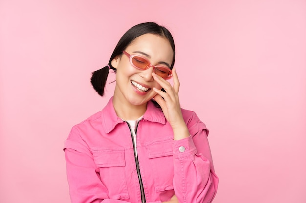 Крупный план портрета стильной азиатки в солнцезащитных очках, улыбающейся, выглядящей польщенной, смеющейся, кокетливой, стоящей на розовом фоне
