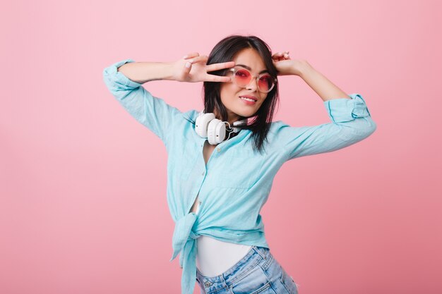 スタイリッシュなアジアの若い女性のクローズアップの肖像画は、エレガントなメガネと綿のシャツを着ています。ピンクの部屋でリラックスした黒い光沢のある髪の愛らしいヒスパニック系の女の子。