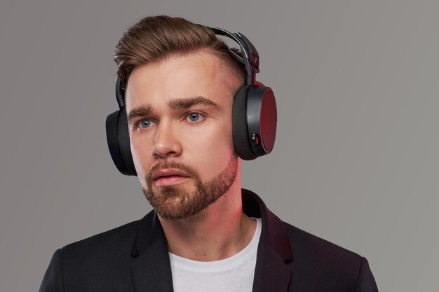 Close-up portrait of a stylish bearded man with hairstyle listening to music in headphones. Isolated on a gray background.