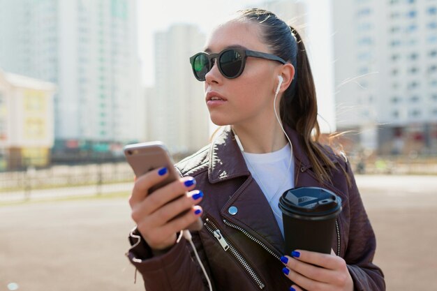 サングラスとジャケットを身に着けている黒髪のスタイリッシュなヨーロッパの女の子の肖像画をクローズアップスマートフォンを持って、街の背景に日光の下で脇を見ています