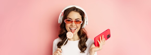 Free photo close up portrait of stylish glamour girl beautiful woman in headphones listening music with smartphone wearing sunglasses and licking lolipop